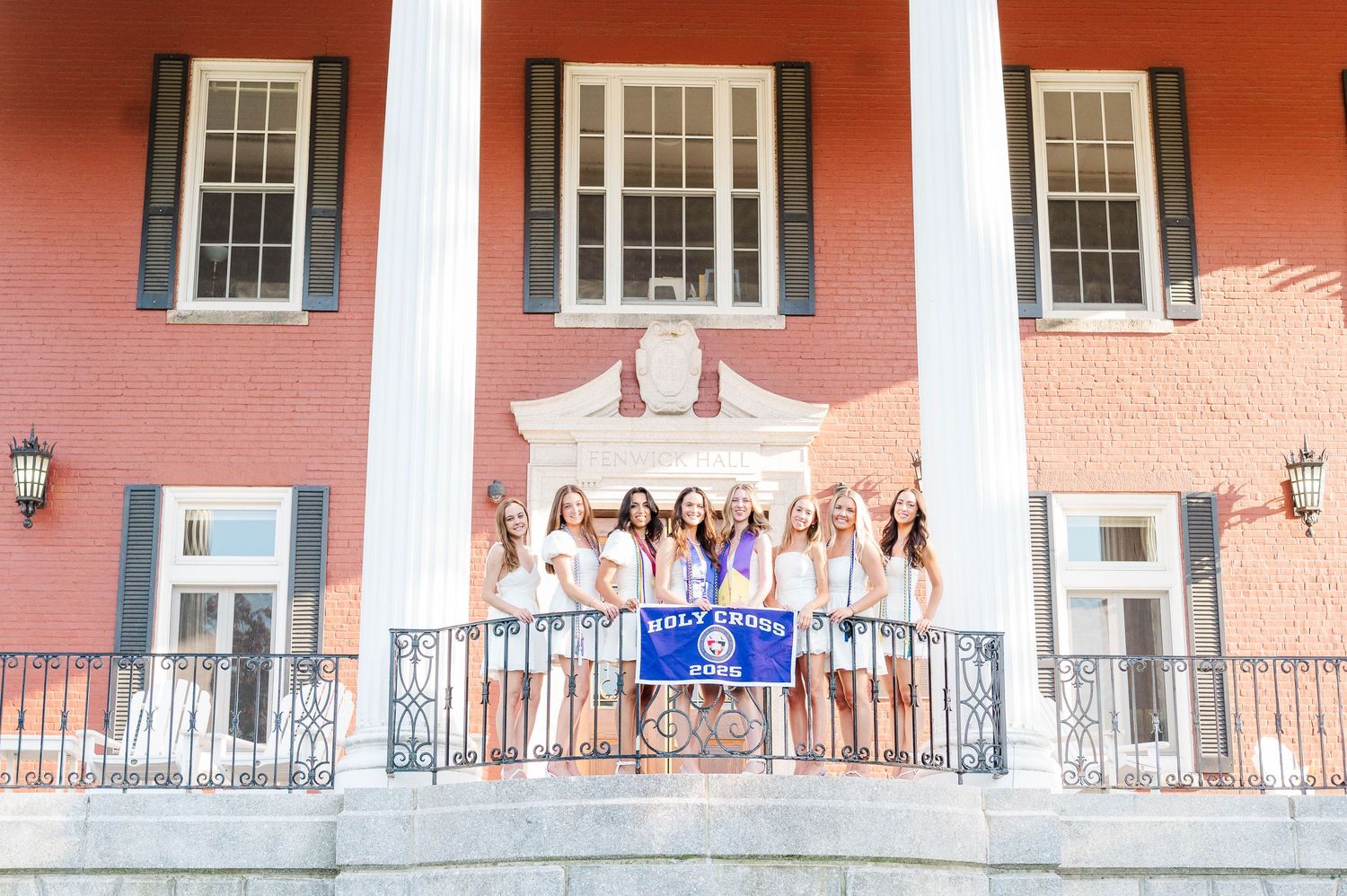 Group of people in white dresses stand on a grand pink porch with white columns at a sorority house.