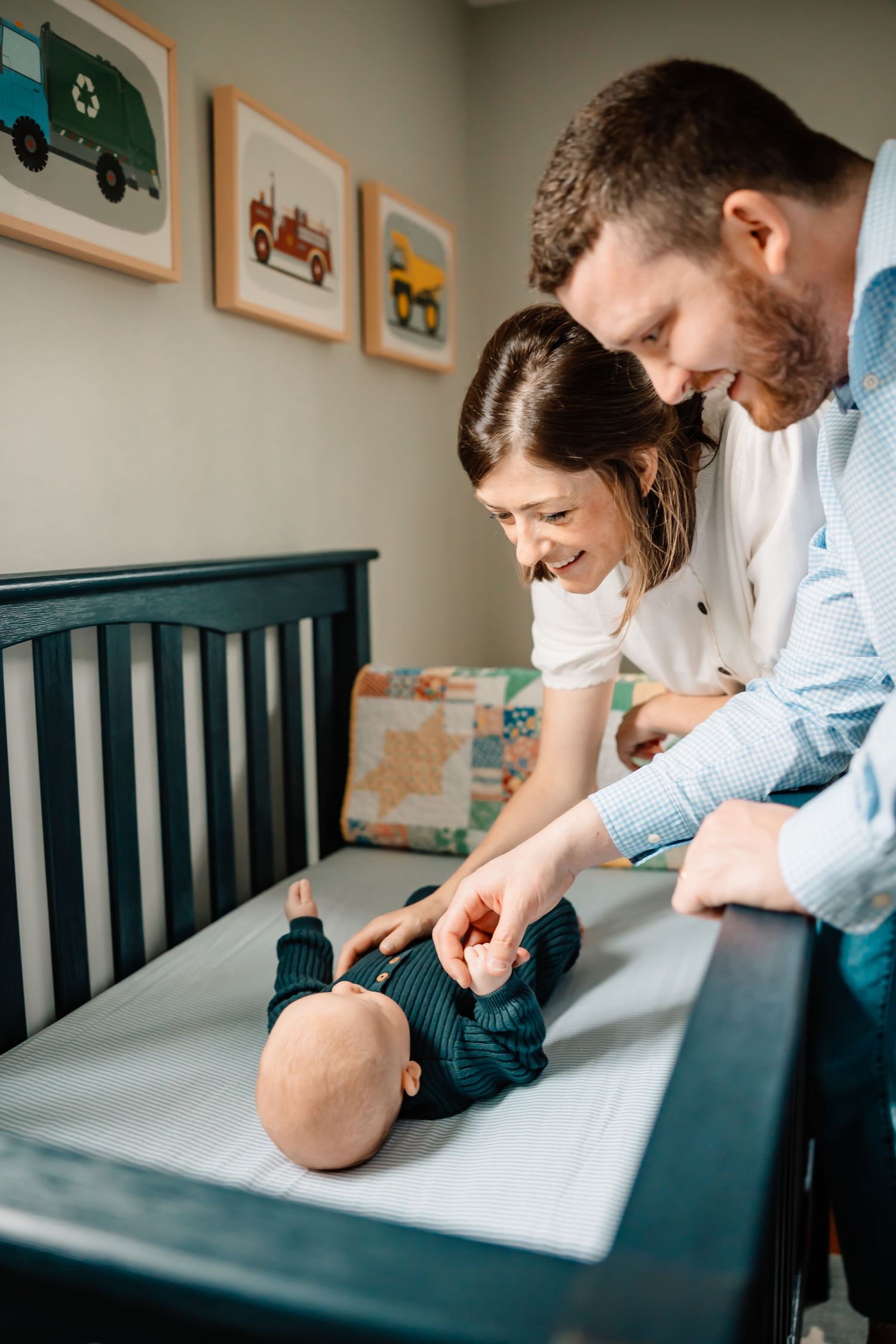 Parents lean over a navy blue crib in a nursery decorated with colorful transportation artwork on the walls.