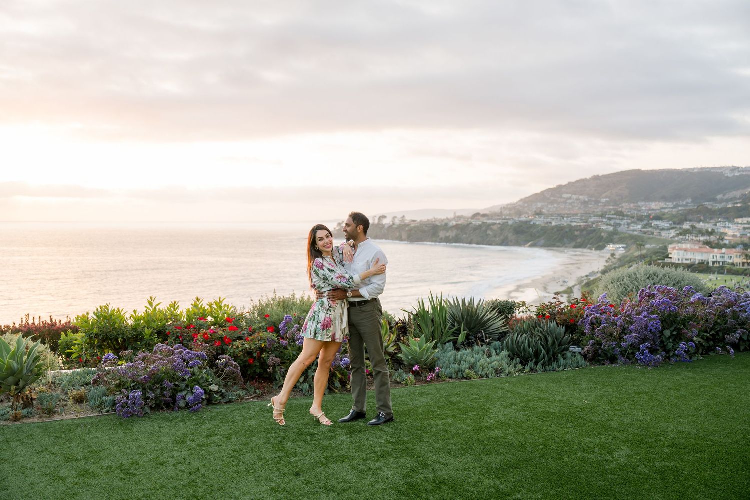 Couple embraces on grassy hilltop overlook with vibrant purple flowers and coastal beach view at sunset.