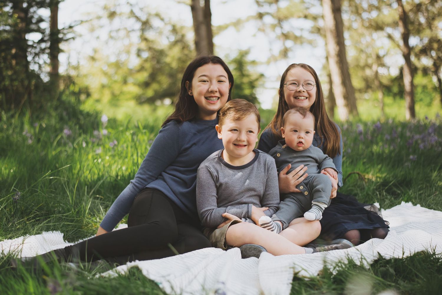 A playful group of siblings sits together on a white blanket in a sunlit woodland setting during a photo session.