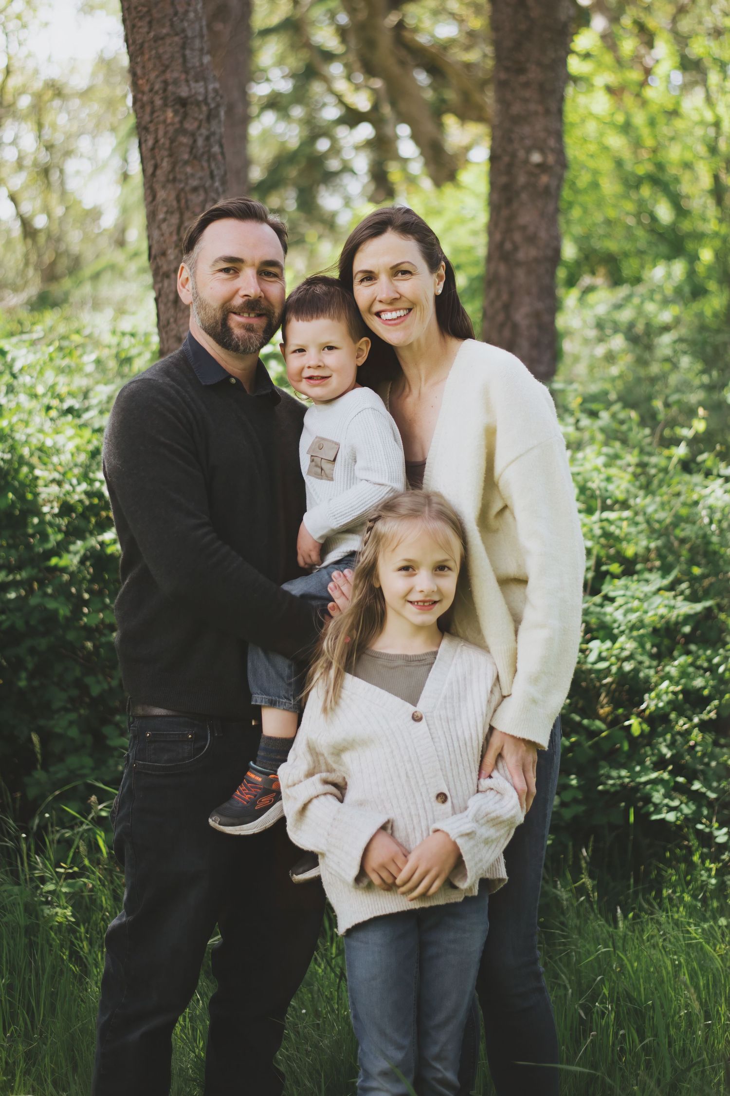 A family stands together outdoors in coordinating cream sweaters against a lush green forest background.