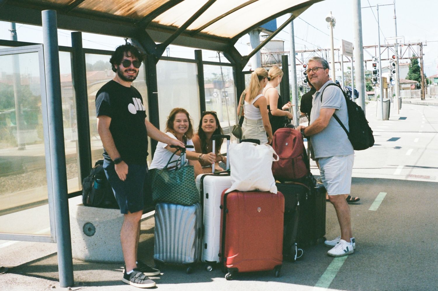 A group of travelers with luggage waiting at a covered bus stop platform on a sunny day.