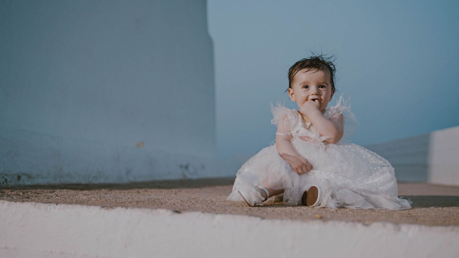 Small child wearing white dress sitting on floor during photo session against blue wall background.