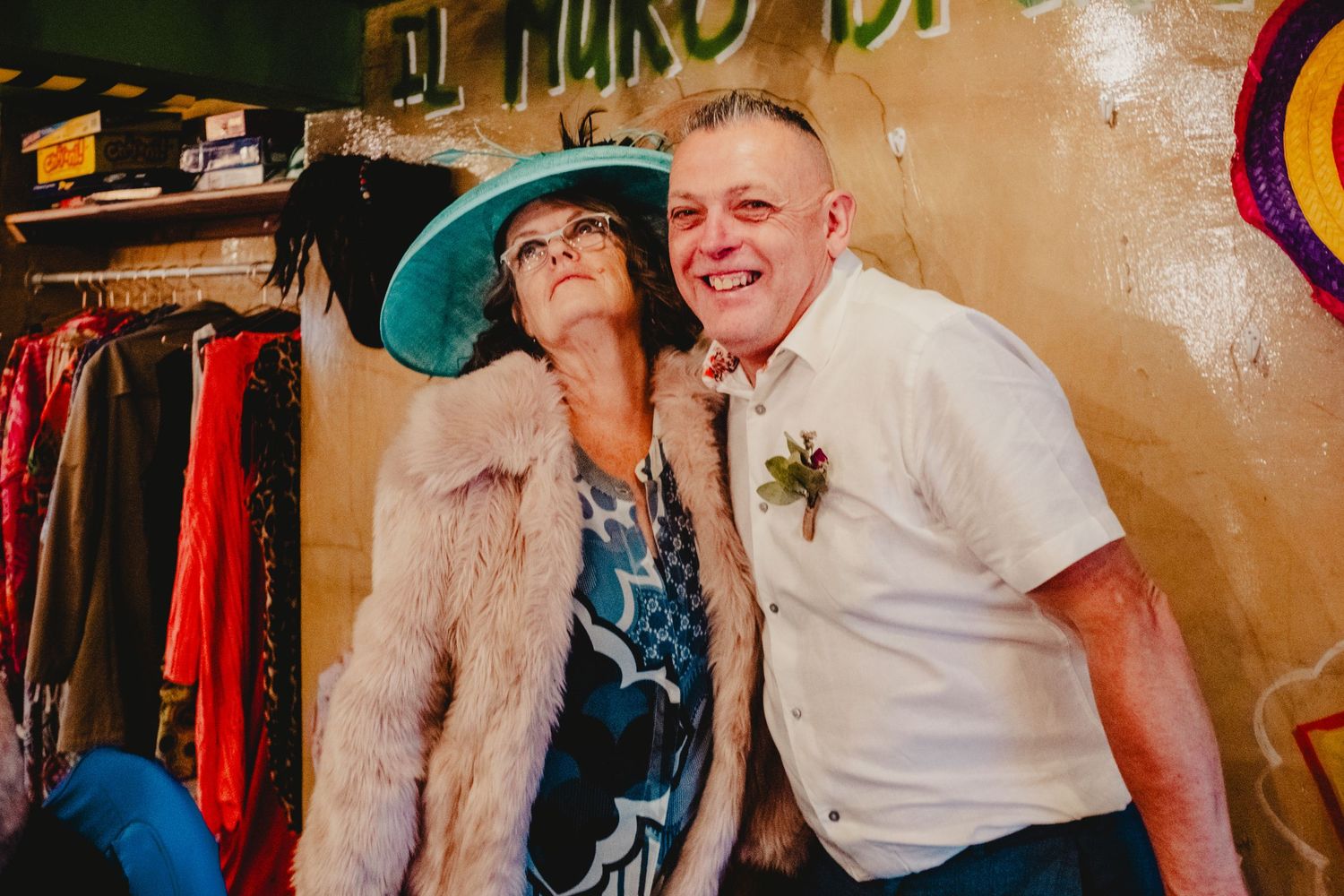 Two people pose playfully together at what appears to be a vintage clothing shop, one wearing a large turquoise hat.