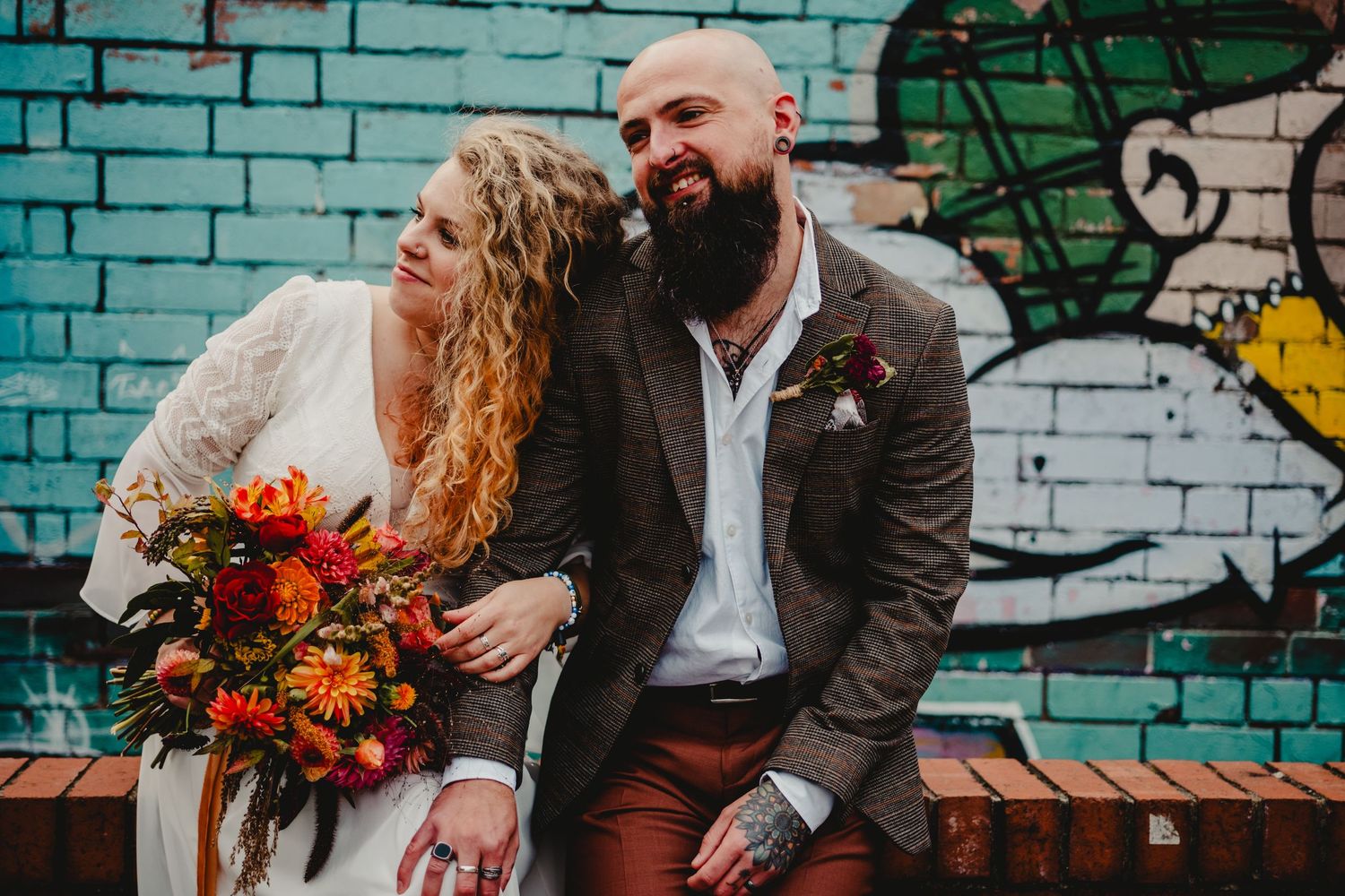 A couple shares a tender moment on a bench against a turquoise graffiti wall, with the bride holding vibrant autumn flowers.