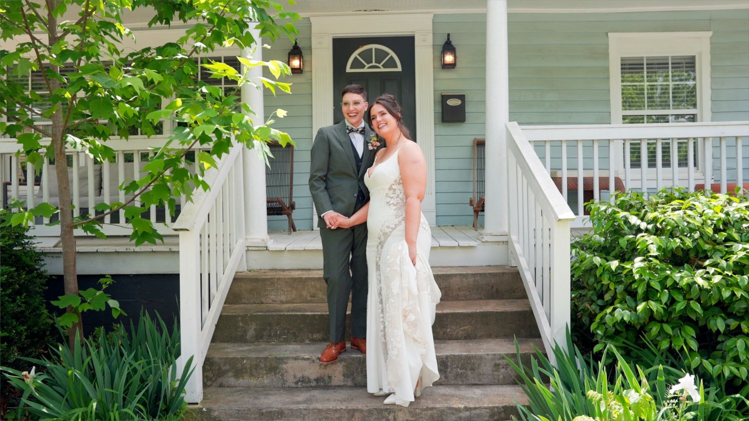A couple stands together on the front porch steps of a charming white house with a green garden.