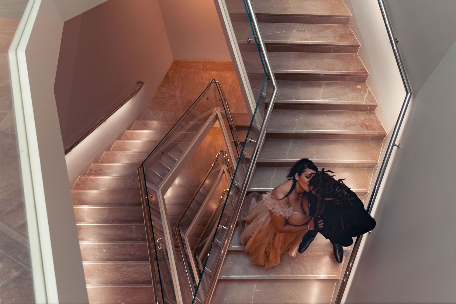 Person in a flowing orange dress sits on elegant wooden staircase with warm lighting and wooden handrails.