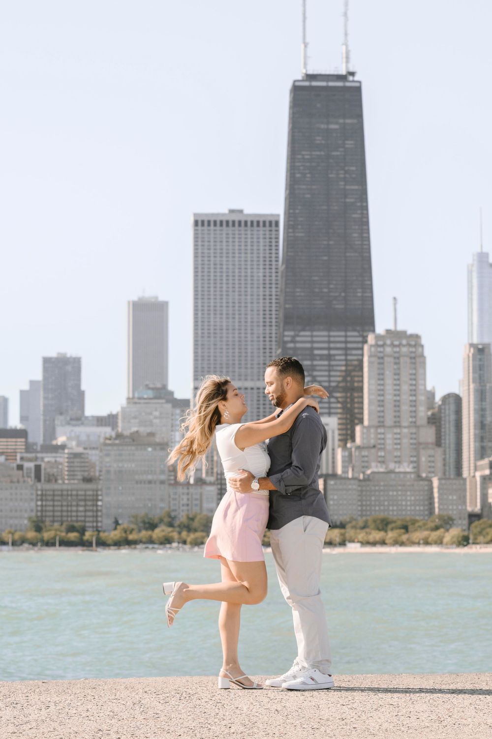 A romantic couple embraces on the Chicago waterfront with the John Hancock building and city skyline in the background.