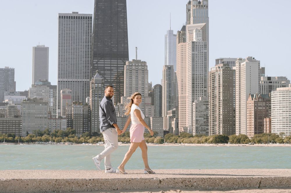 Two people walk along Lake Michigan with Chicago's iconic skyline of tall skyscrapers in the background.