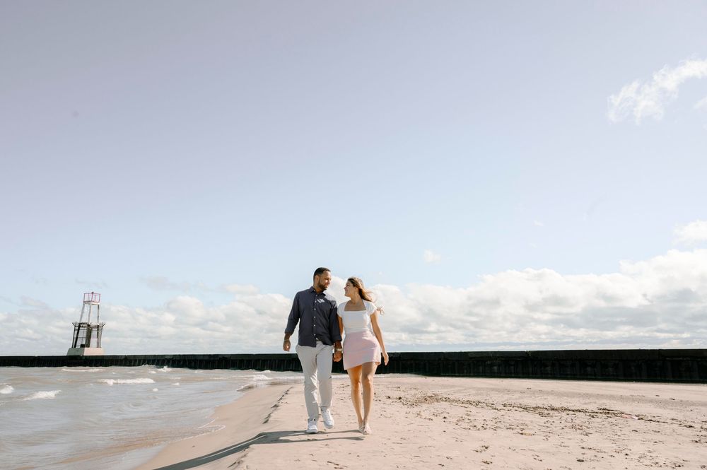 A couple walks together along a sandy beach shoreline with a breakwater and lighthouse in the distance.