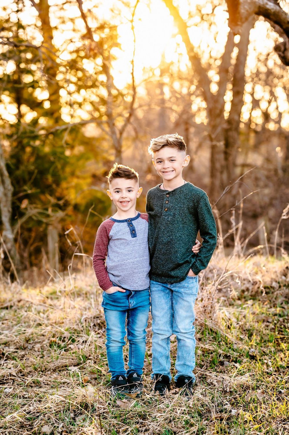 Two children stand together in casual sweaters and jeans against a golden sunset in a wooded outdoor setting.