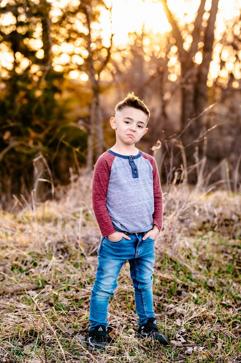 Child in grey and red raglan shirt and blue jeans poses outdoors during golden hour sunset in a wooded area.
