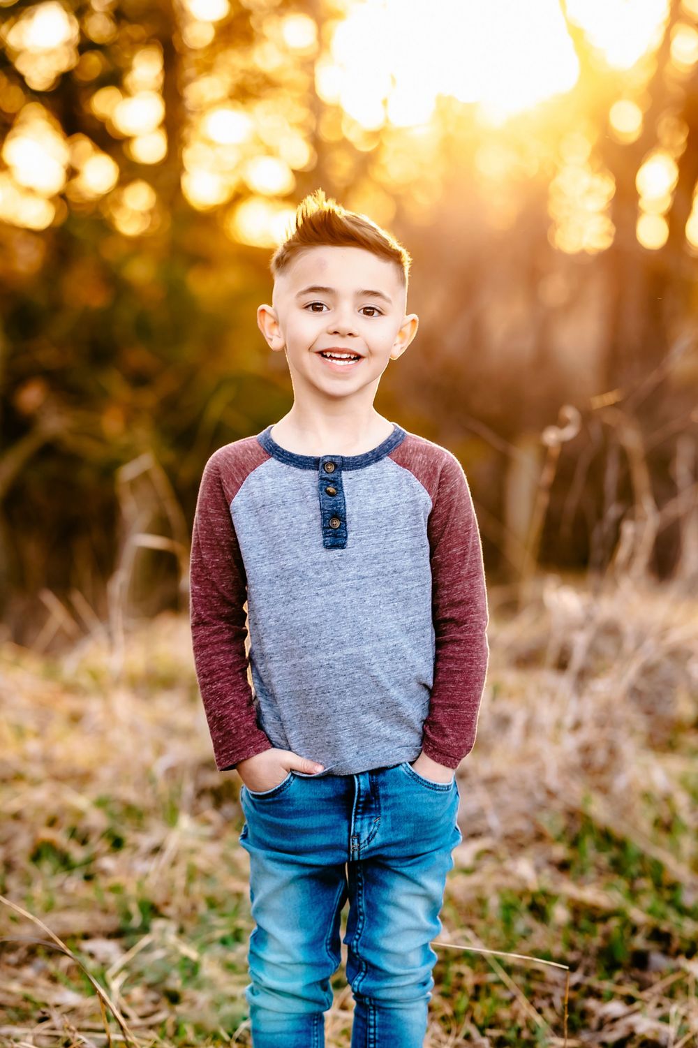 A portrait of a child wearing a grey and burgundy raglan shirt against a warm glowing forest backdrop.