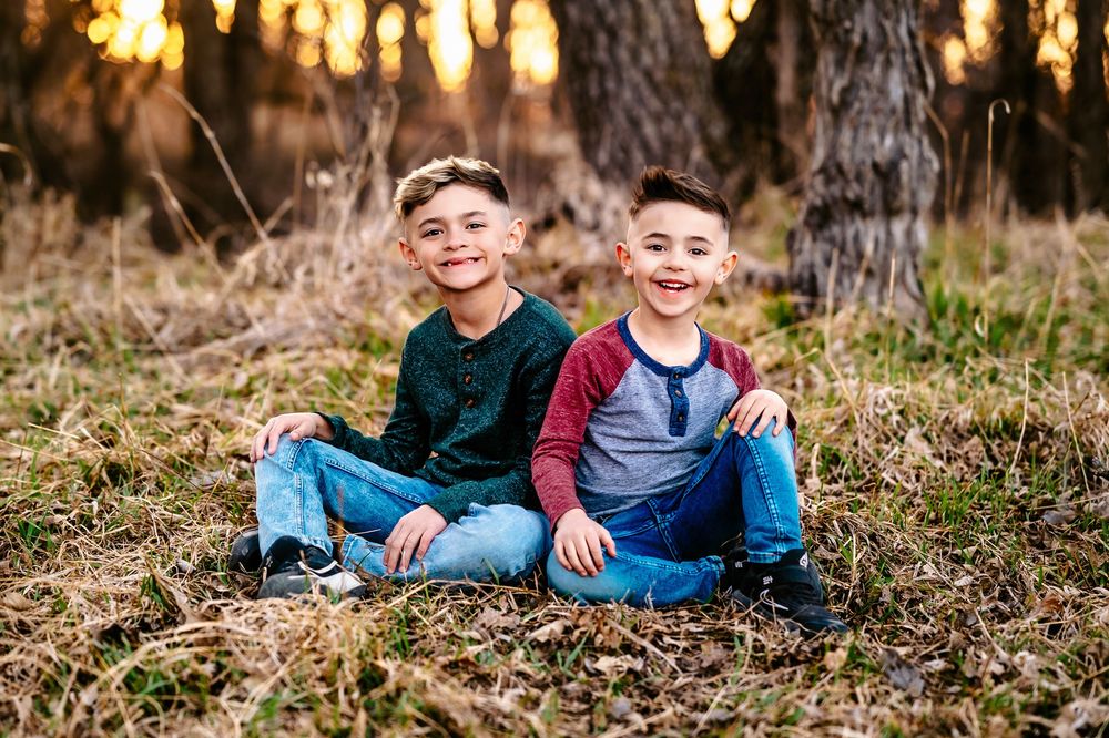Two children sitting side by side on autumn ground with warm sunset lighting through trees behind them.