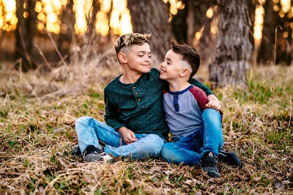 Two young boys sitting together and laughing on fallen leaves during sunset in a wooded area.