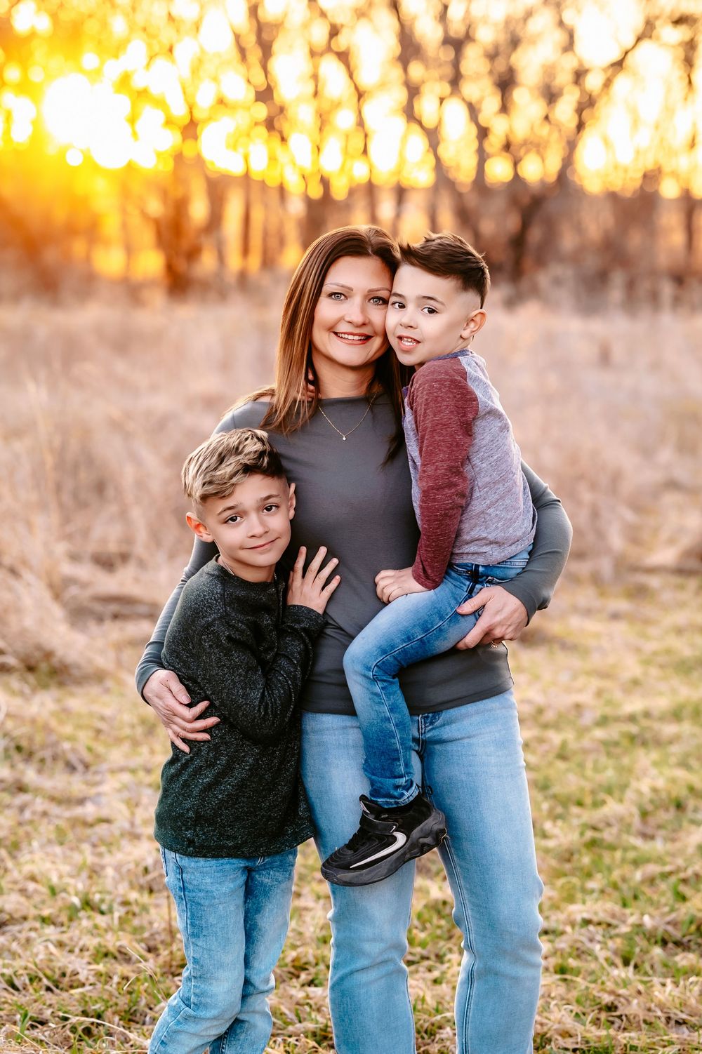Family portrait of three people outdoors with golden sunset light filtering through trees in background.