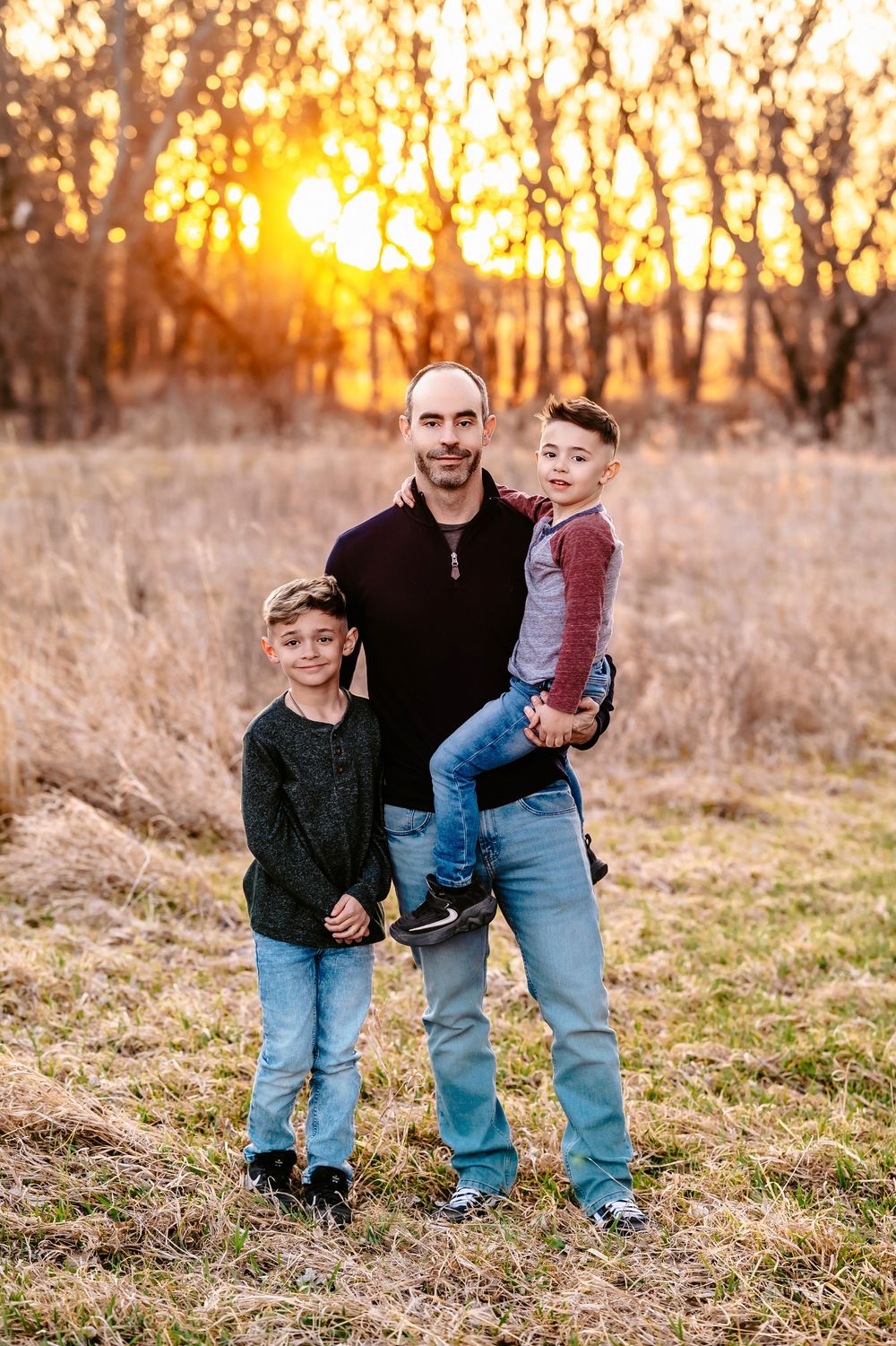 Three people posing for a family portrait at sunset with golden light filtering through trees behind them.