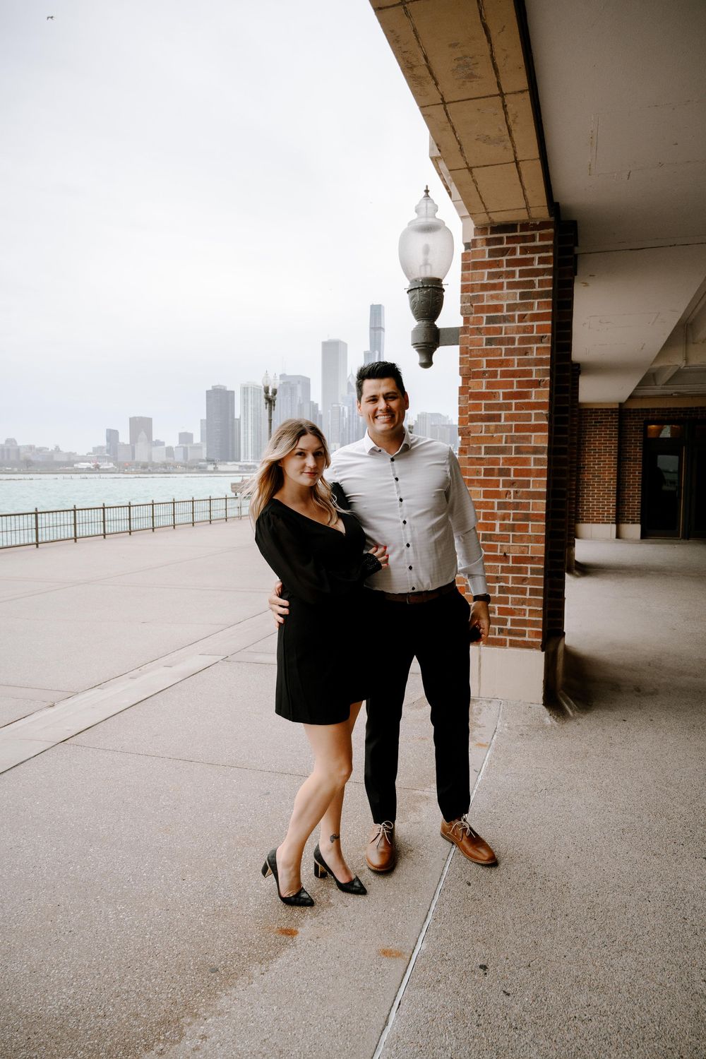 Couple poses together on urban waterfront promenade with city skyline visible across the water on overcast day.