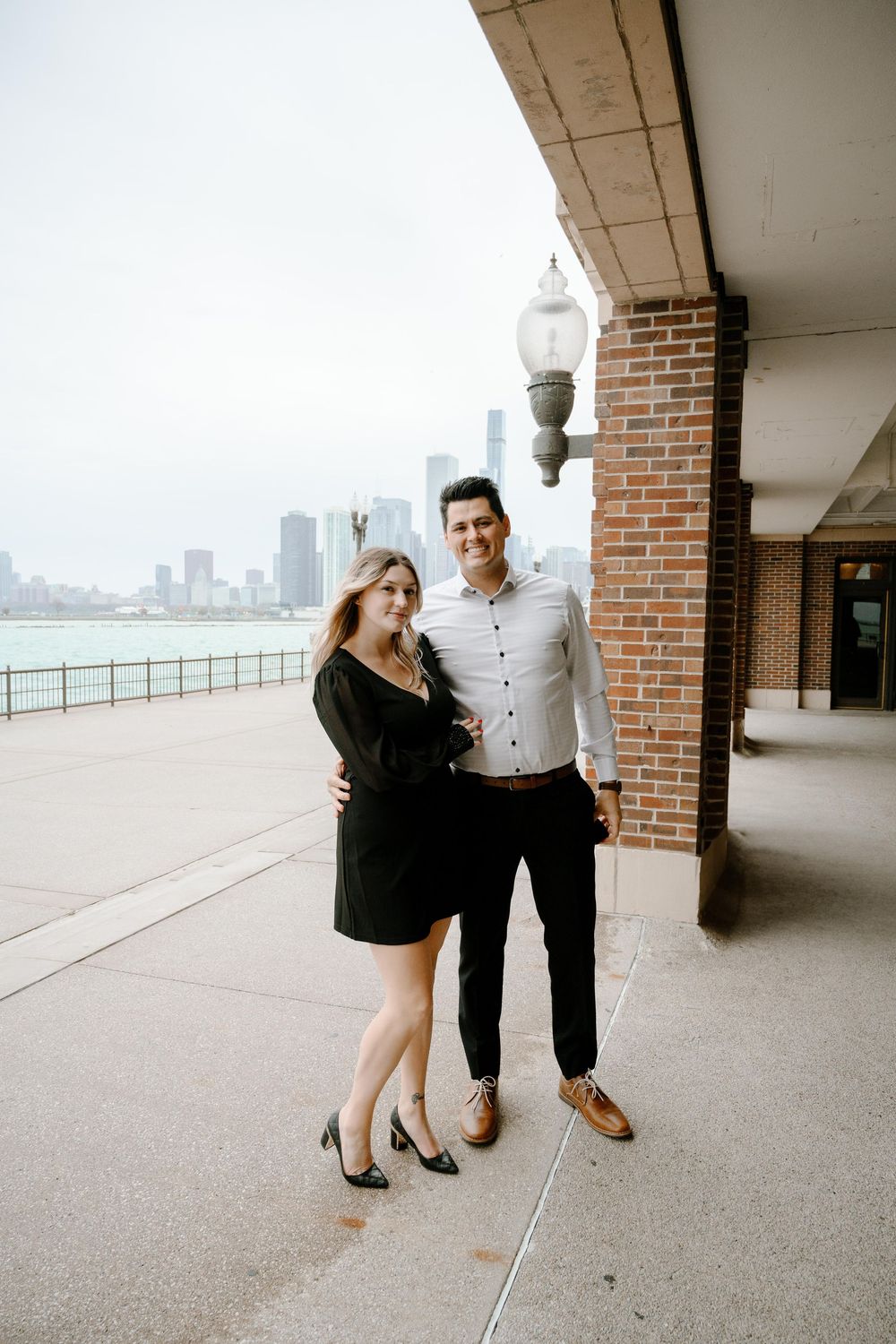 Couple stands together on scenic waterfront walkway with Chicago skyline in background on cloudy day.