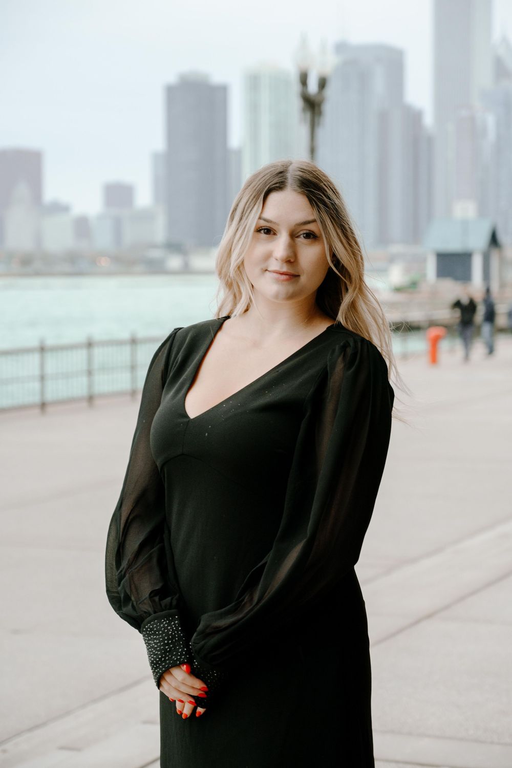 A woman in a black dress poses on a waterfront promenade with city skyscrapers in the background.