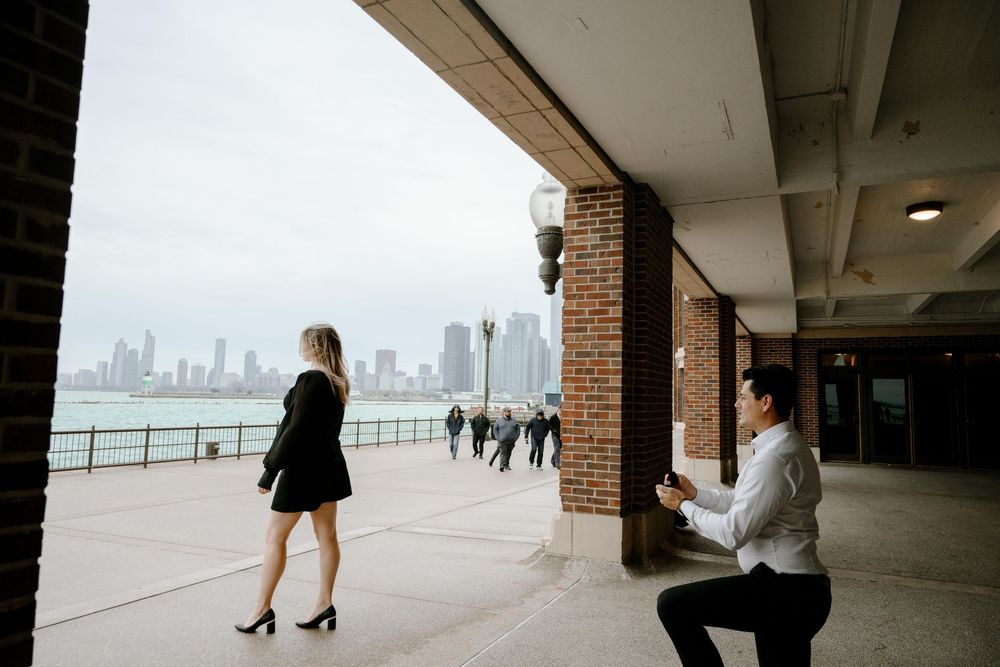 A person kneels on one knee under a covered walkway along a waterfront with city skyline visible in the distance.
