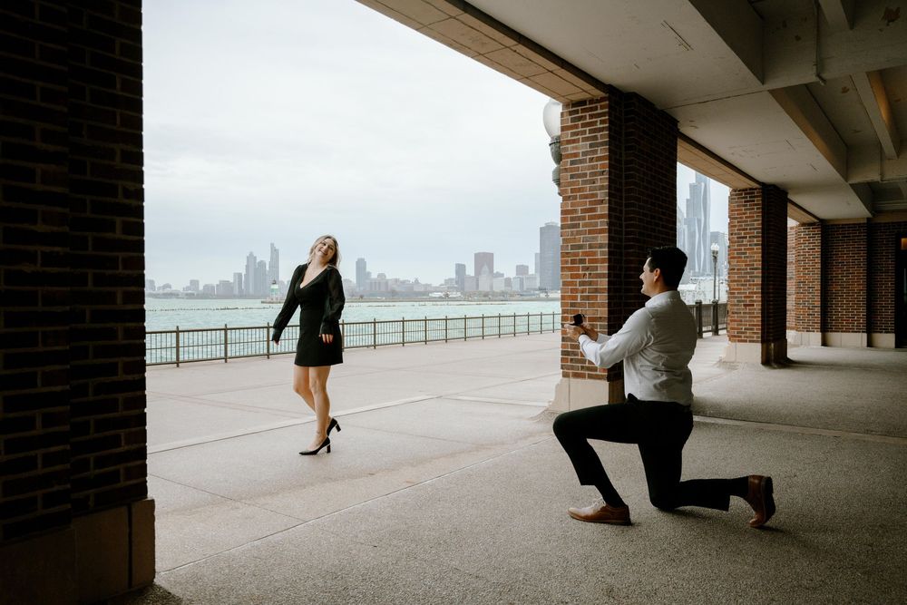 A romantic proposal moment at a covered walkway overlooking a city skyline across the water.