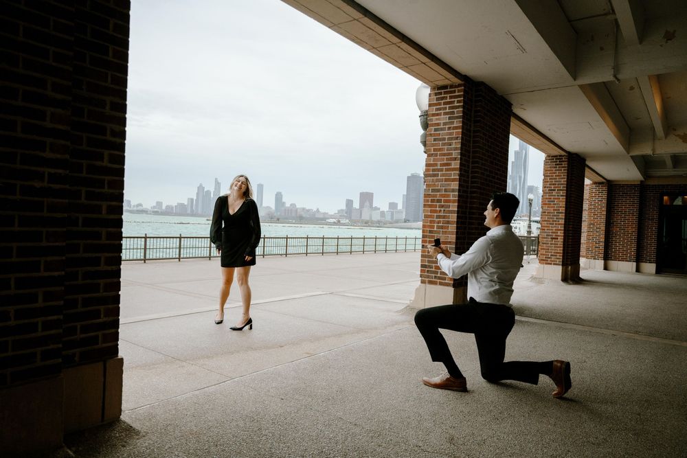 A couple shares a romantic proposal moment under a covered walkway with a cityscape view in the background.
