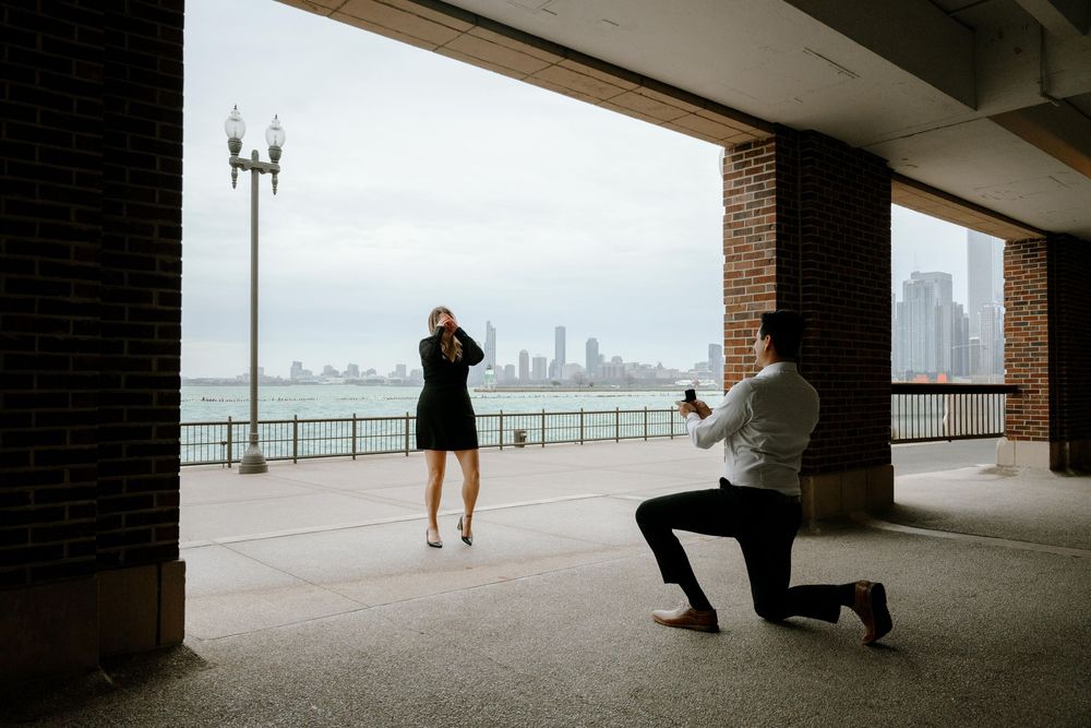 A couple shares a romantic proposal moment with Chicago skyline in view at a covered waterfront location.