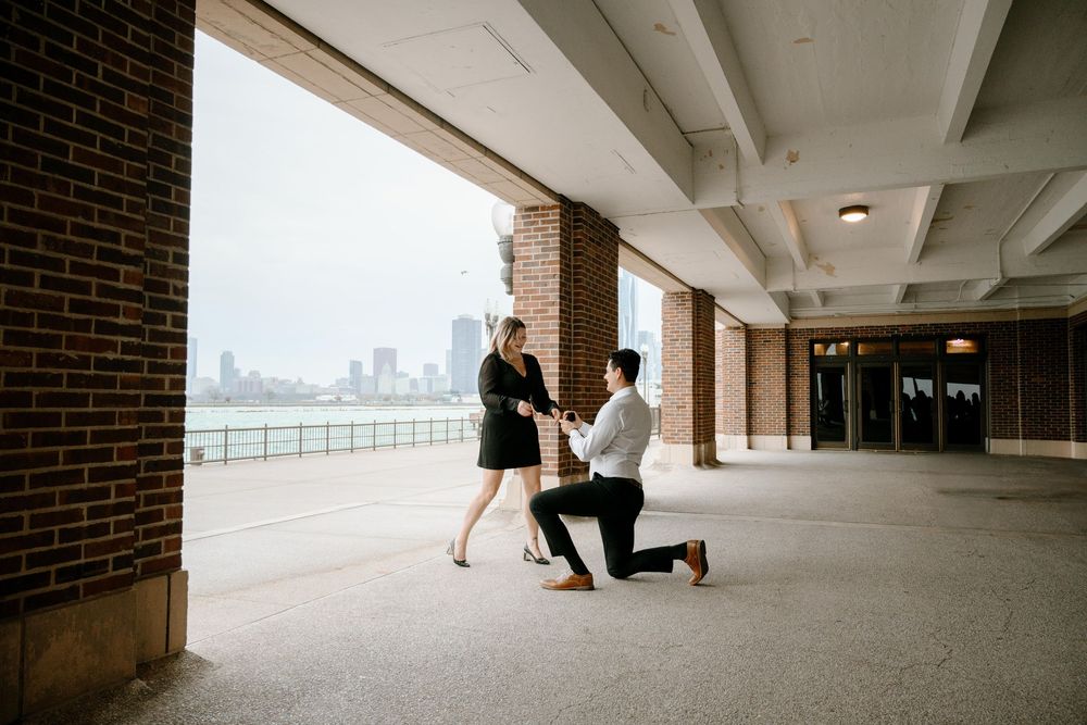 A romantic proposal moment between a couple at a covered brick colonnade overlooking a city skyline.