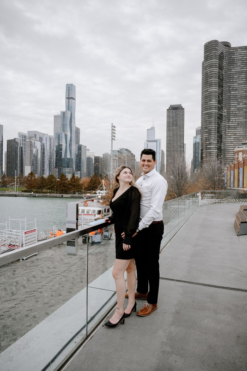 A couple poses along the Chicago Riverwalk with a backdrop of modern skyscrapers on a cloudy day.