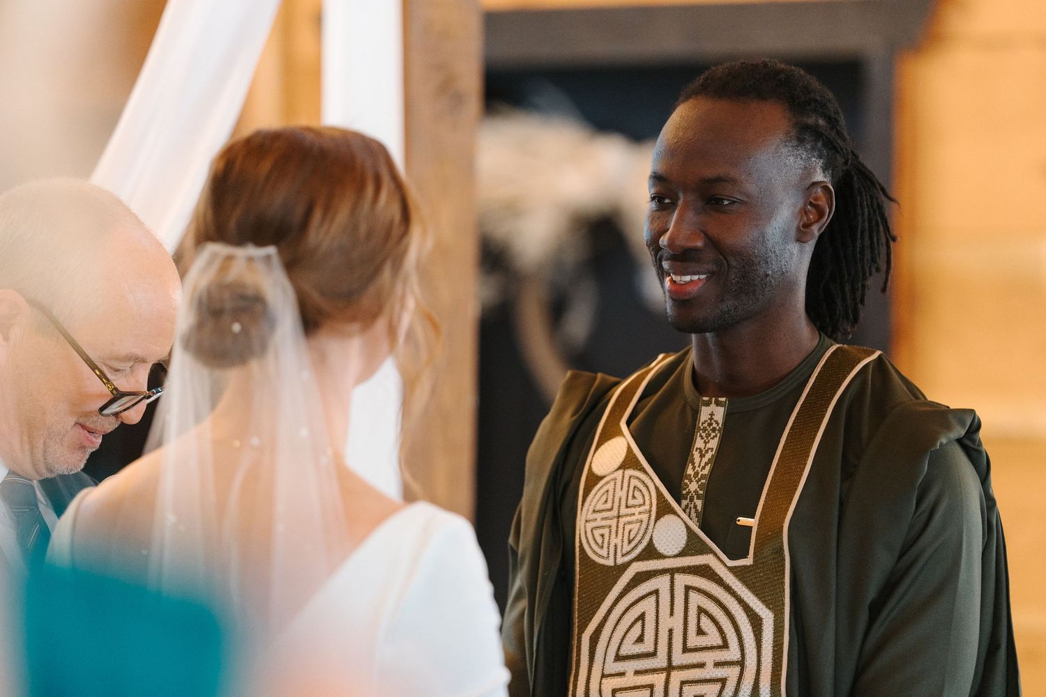 A joyful wedding moment shows a person in traditional dress with patterned symbols smiling at someone in a white veil.