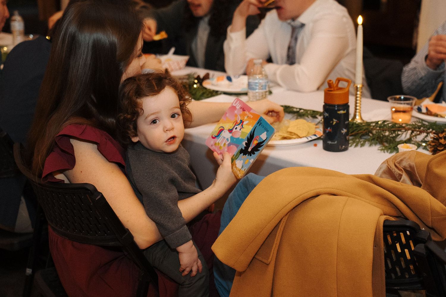Child seated at restaurant dinner table playing with a blue toy while surrounded by plates of food.
