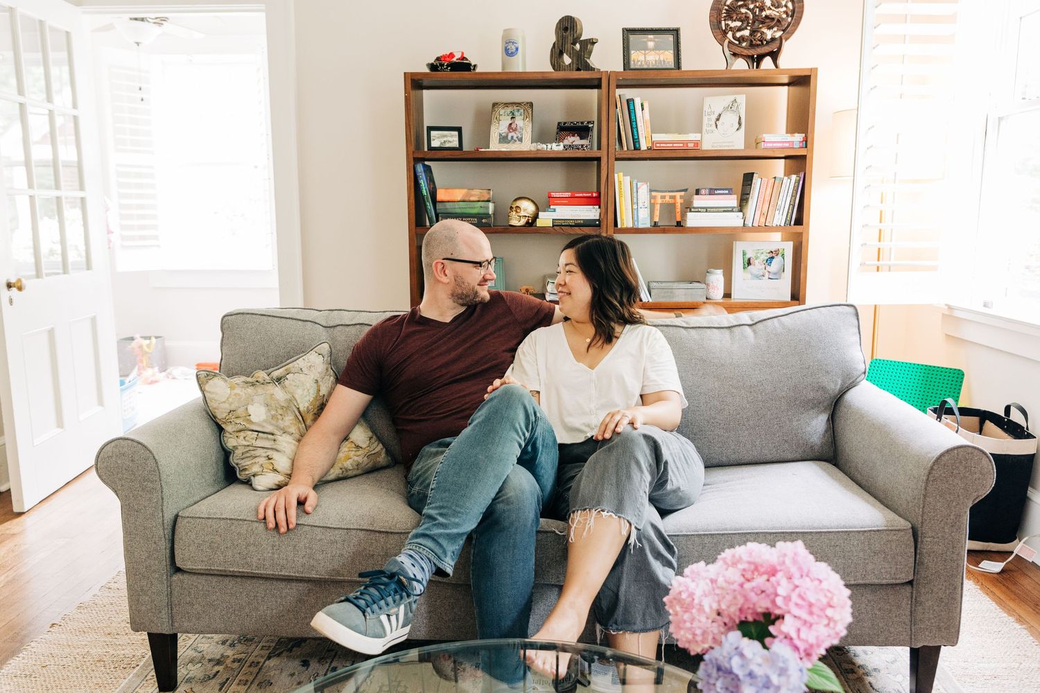 A couple relaxes together on a gray couch in a bright living room with wooden shelves and decor in the background.