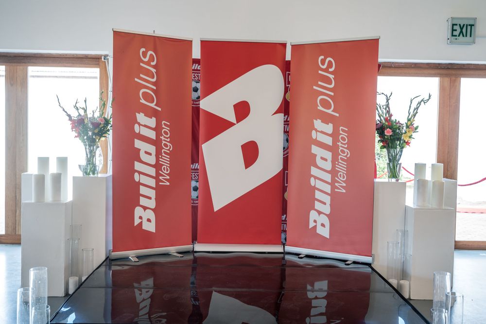 Three red Buildprix banners displayed in an indoor venue with exit sign visible.