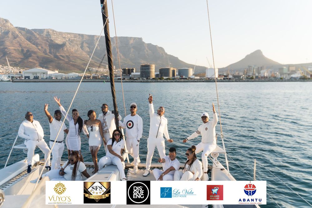 A group in white attire celebrating on a yacht with Table Mountain visible in Cape Town's harbor.