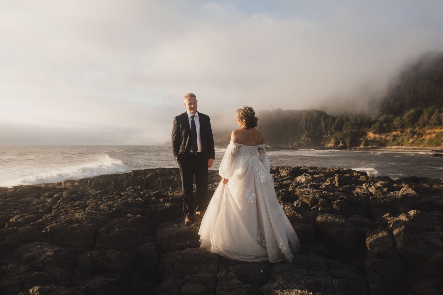 A scenic wedding photo on a misty rocky beach with cliffs and ocean waves in the background.