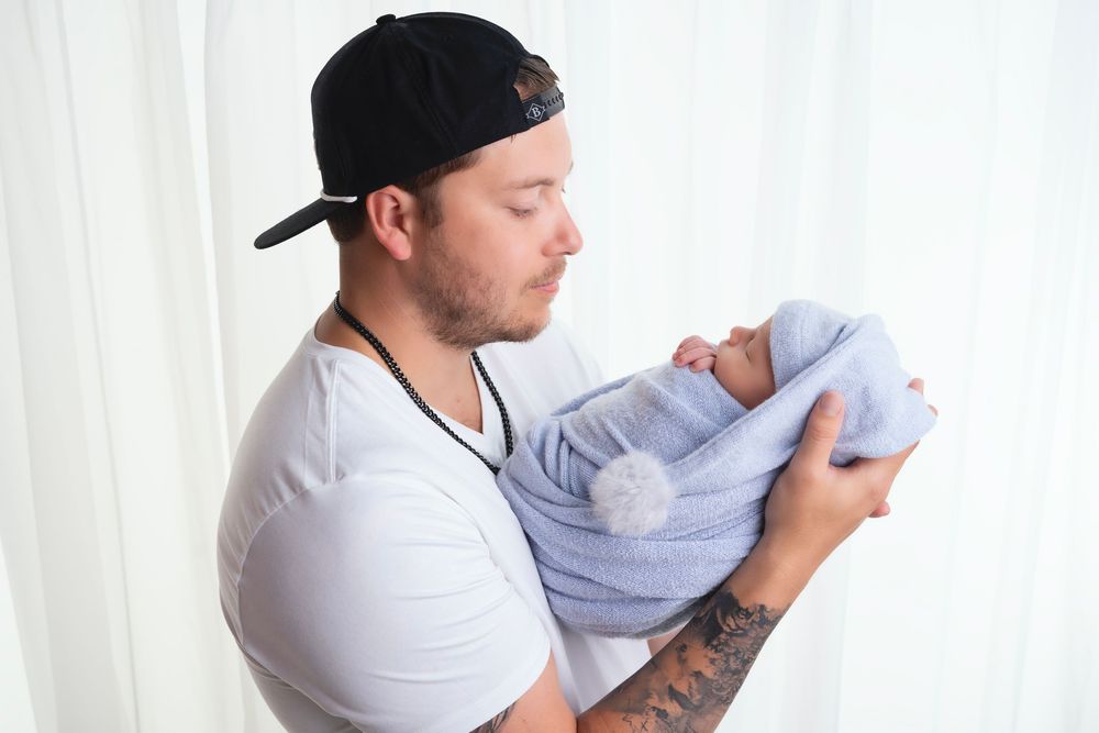 A person in a white shirt and baseball cap cradles a sleeping newborn wrapped in a lavender blanket.