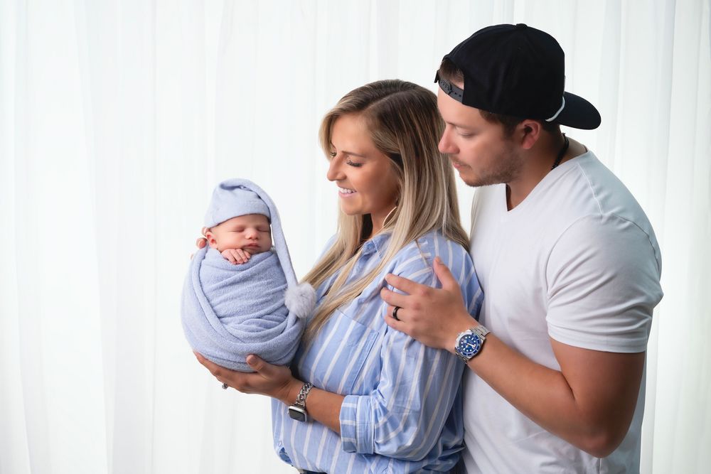 A couple in matching blue and white outfits tenderly hold their swaddled newborn baby against a white curtain background.