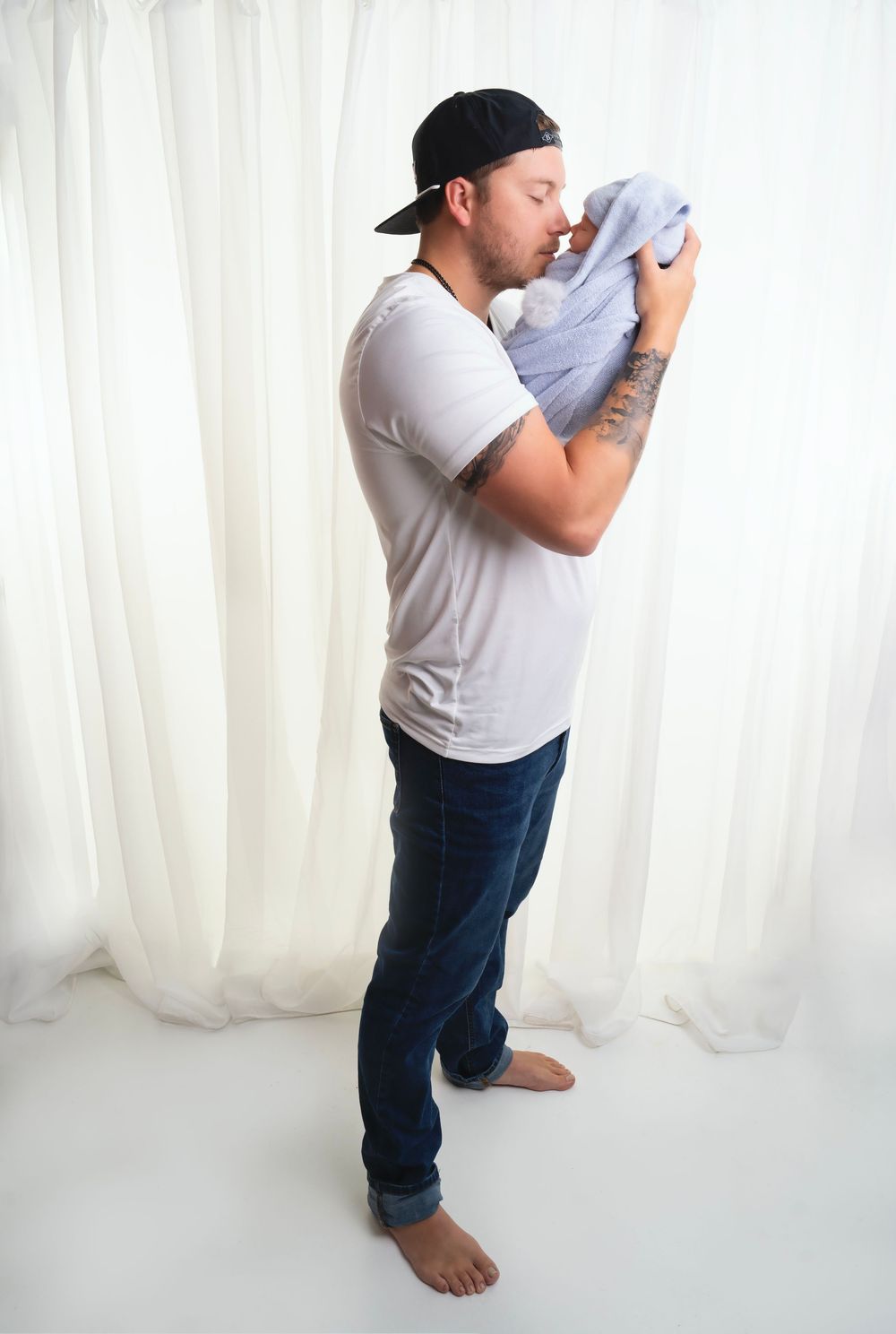 Side view of person in white t-shirt, jeans and baseball cap holding something soft against their face while standing barefoot.