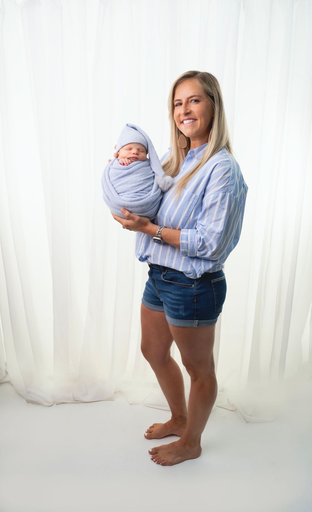 Mother in striped shirt and denim shorts stands barefoot holding swaddled newborn baby against white backdrop.