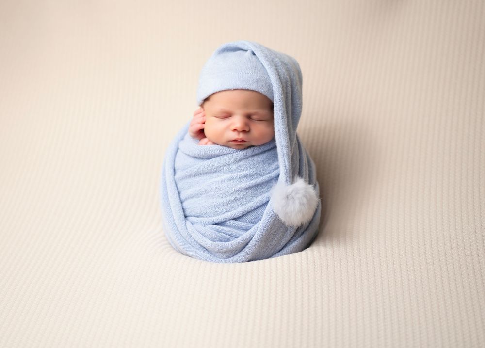 A newborn baby wrapped in soft blue blankets with a matching hat rests peacefully against a beige backdrop.