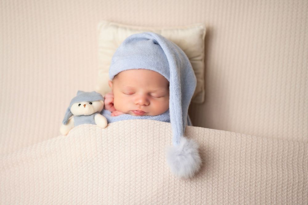 A sleeping baby wearing a light blue knit hat rests on cream bedding with a small plush toy companion.