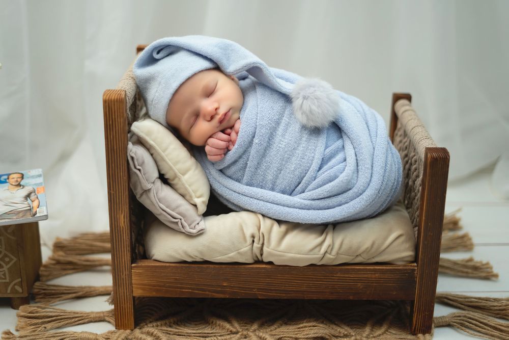 Tiny newborn wrapped in soft blue swaddle sleeping peacefully on cushioned wooden bed.