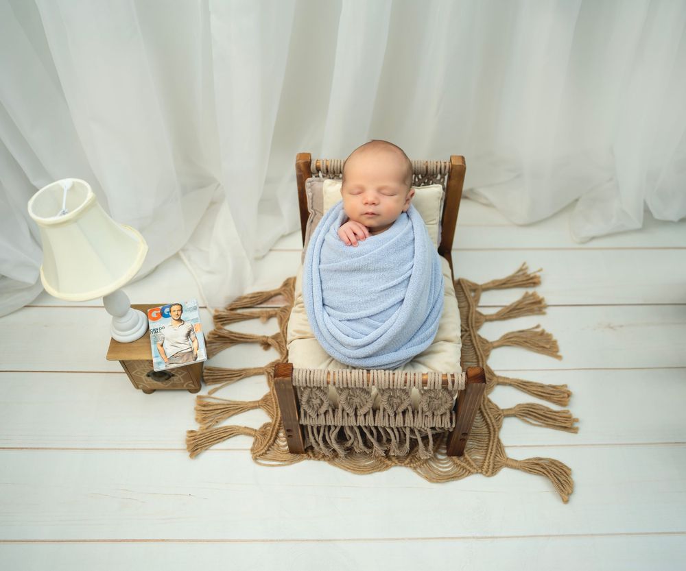 A sleeping baby wrapped in blue cloth lies on a brown fringed blanket in a wooden chair with a lamp nearby.