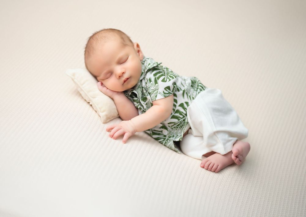 Peacefully sleeping baby wearing floral shirt and white pants rests on textured beige blanket in studio setting.
