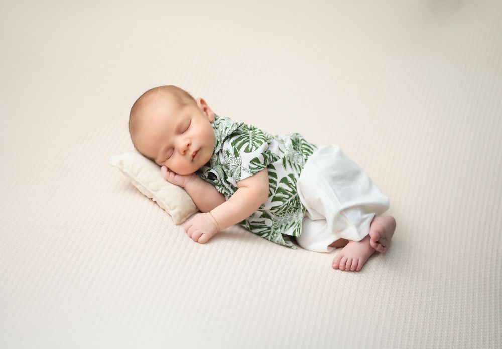 A sleeping baby wearing a green floral outfit rests peacefully on a cream-colored textured surface.