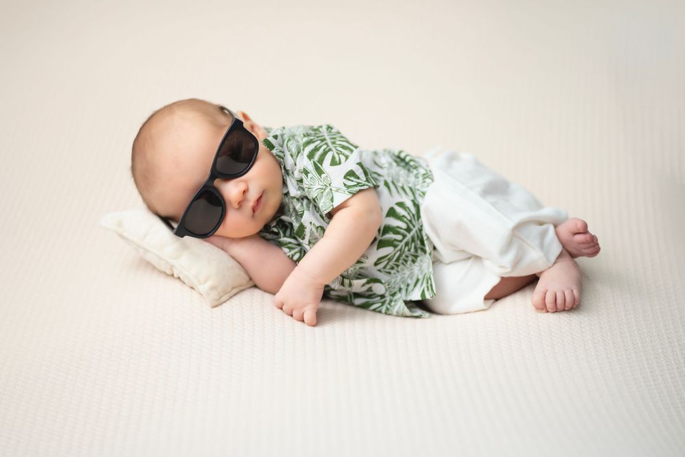 A baby wearing dark sunglasses and a tropical print shirt relaxes on a white pillow.