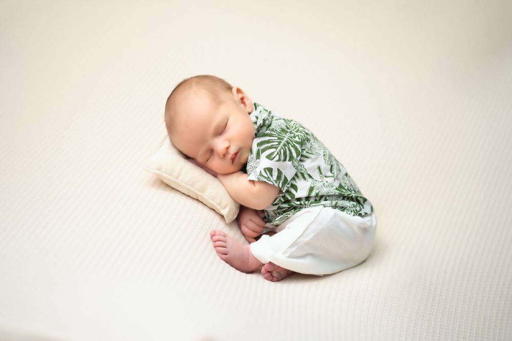 A sleeping baby wearing a white and green leaf-patterned outfit rests on a cream-colored cushion.