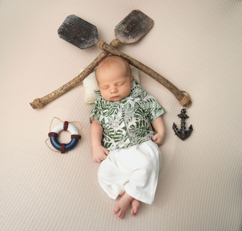 A sleeping baby in tropical print lies beneath crossed wooden oars with nautical decorations on beige fabric.