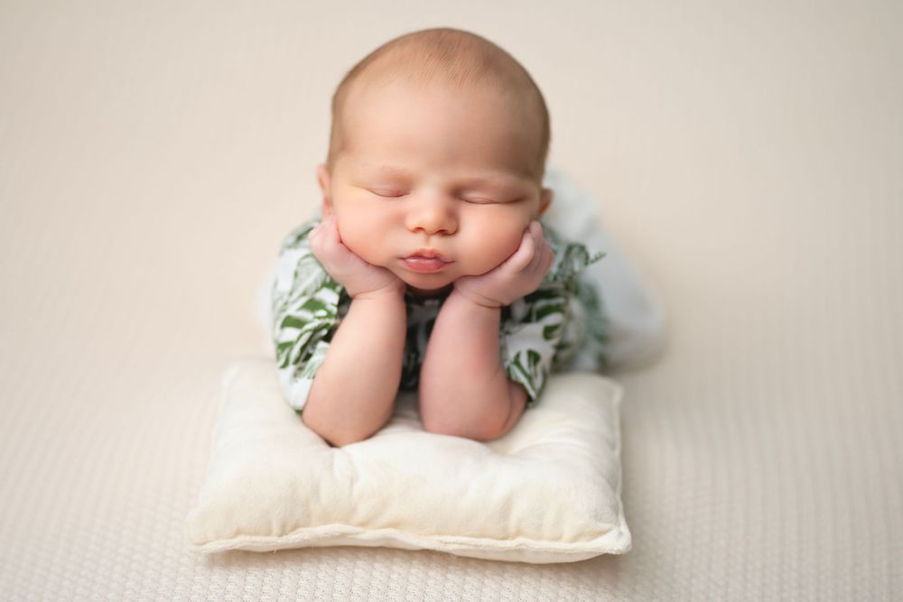 Sleeping newborn resting head on hands while lying on white pillow wearing mint green outfit in studio setting.